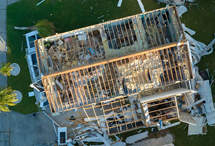 Aerial view of a house with its roof completely destroyed by a storm. Debris litters the surrounding lawn.