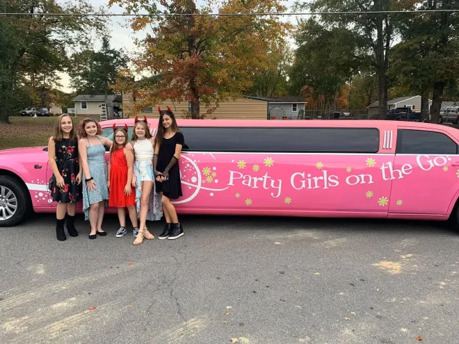Group posing beside pink limousine reading “Party Girls on the Go” in a parking lot.
