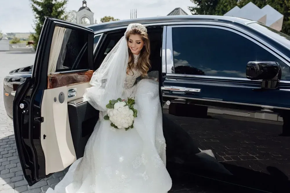 Bride in a white gown and veil stepping out of a black limousine, holding a bouquet.