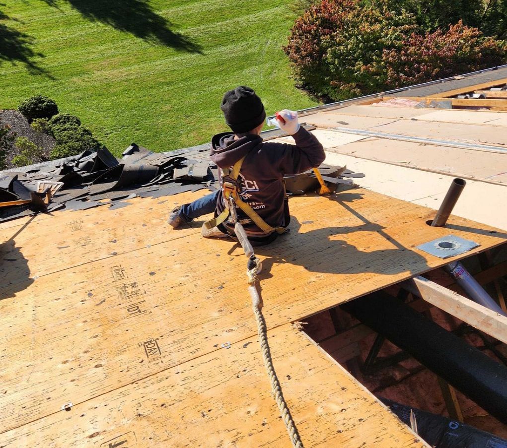 A person in a harness and safety rope sits on a partially shingled residential roof.