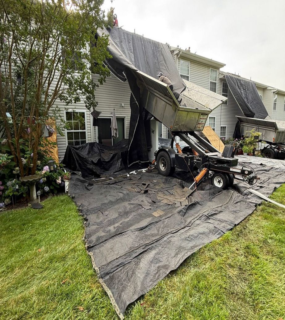 A mechanical lift dumps old roof shingles and debris from a residential townhouse roof onto a large protective tarp below.