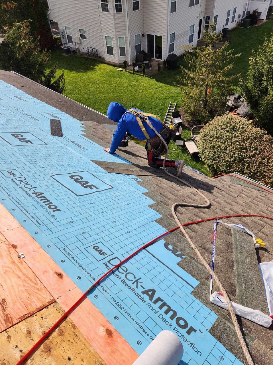 A person in a blue hoodie and safety harness installs blue roof underlayment on a residential roof.