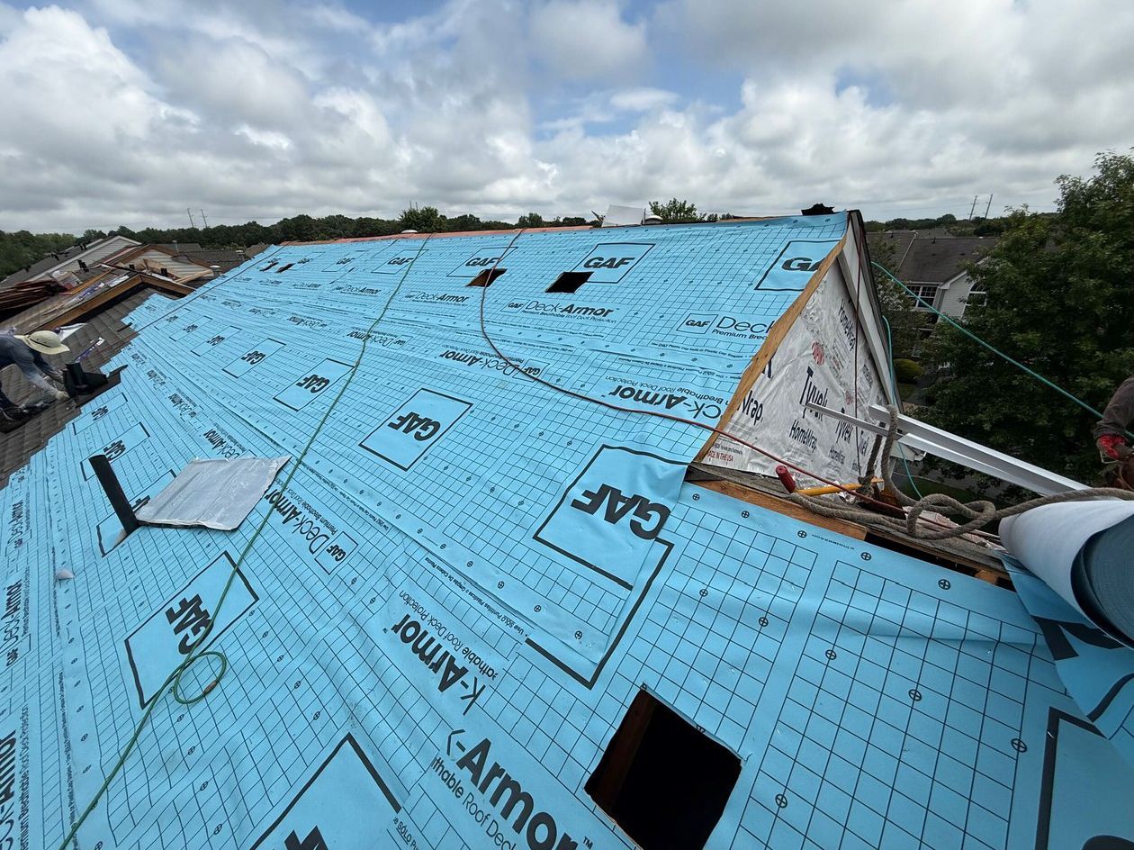 A roof under construction covered in blue GAF brand underlayment, with exposed wood framing and openings for vents.