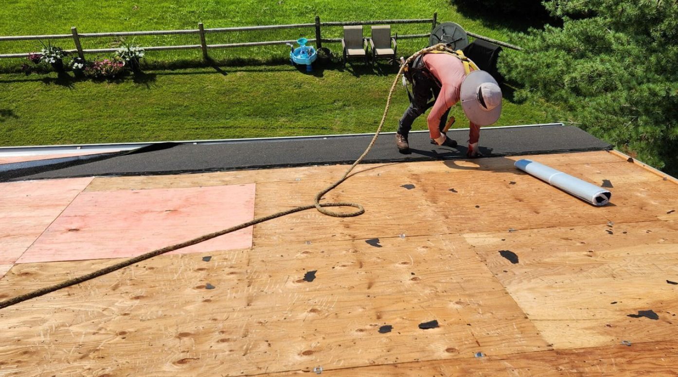 A person in a sun hat and safety harness installs black roofing underlayment on an unfinished plywood roof.