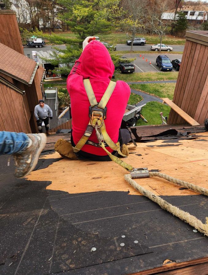 A person in a red hoodie and safety harness sits on a partially shingled roof, with another person visible below.