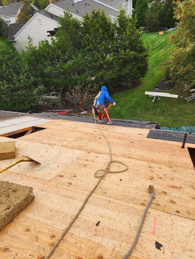 A worker in a blue hoodie on a partially shingled roof, attached to a safety rope, with a grassy yard in the background.