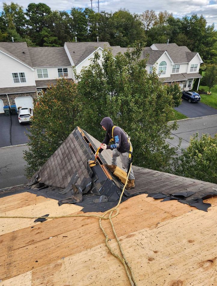 A worker in a gray hoodie and safety harness removes old shingles from a steep residential roof.