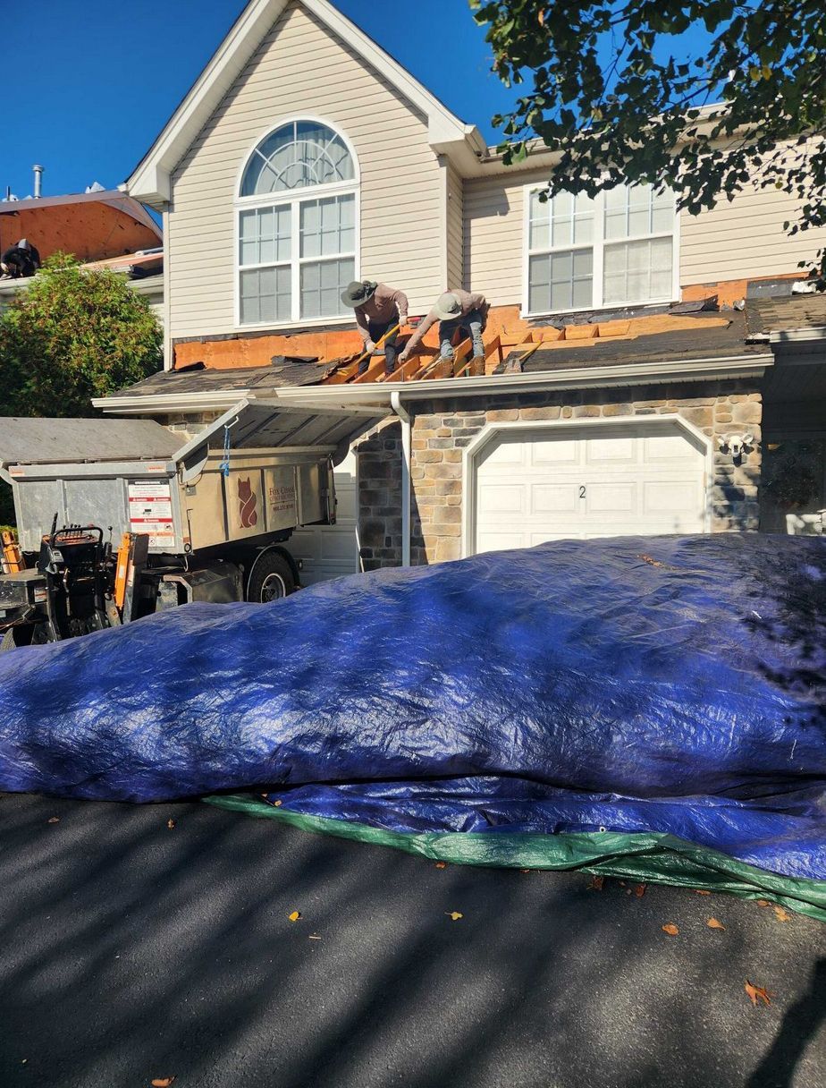 Two workers in hard hats remove shingles from a residential roof while a large blue tarp covers the driveway below.
