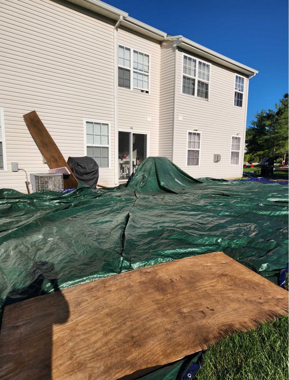 Exterior view of a beige two-story house with a large green tarp covering the backyard ground and wooden boards nearby.