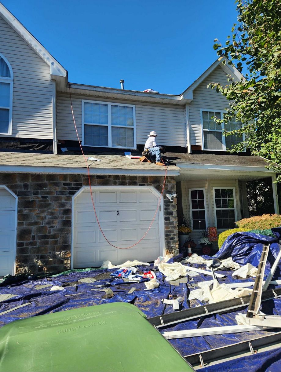 A worker sits on the roof of a two-story home during a roofing project, with construction debris on the lawn below.
