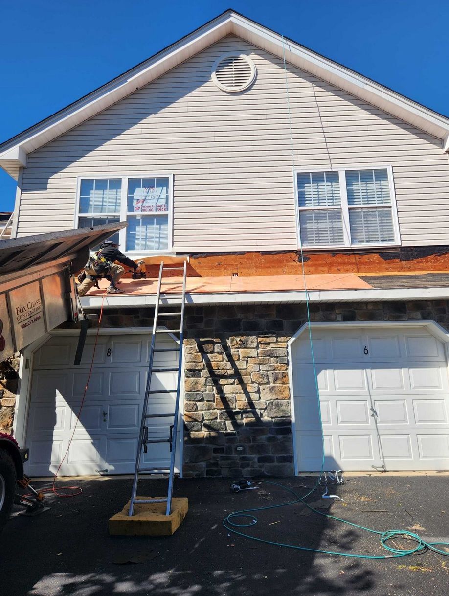 A worker on a ladder performs repairs on the roof above two garage doors on a two-story home with siding and stone facade.