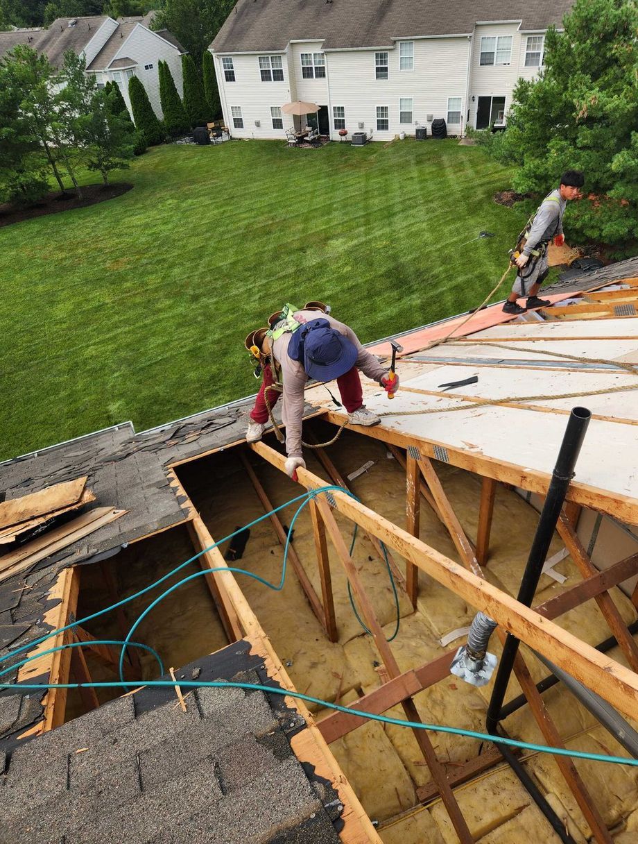 Two construction workers repair an open roof section on a house, visible from an elevated angle above a green lawn.
