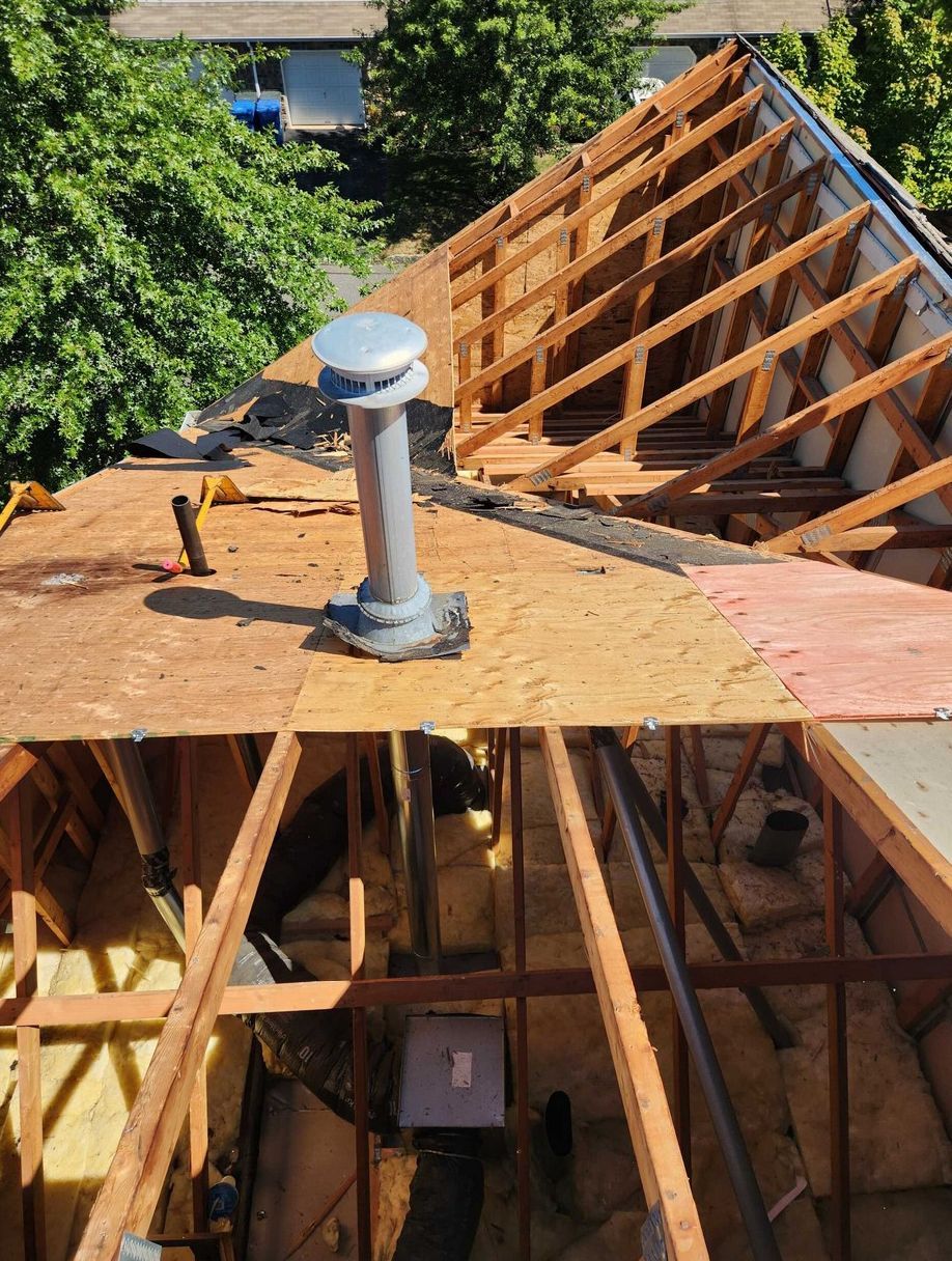A high-angle view of a residential roof under construction with exposed wooden rafters, plywood decking, and a metal vent.
