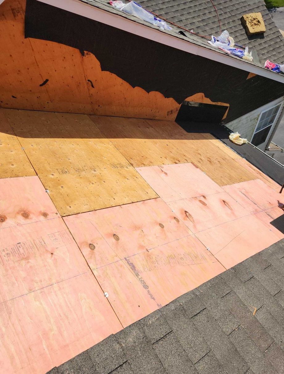 An unfinished roof repair showing a mix of new pink-tinted and light wood panels installed over the existing shingles.