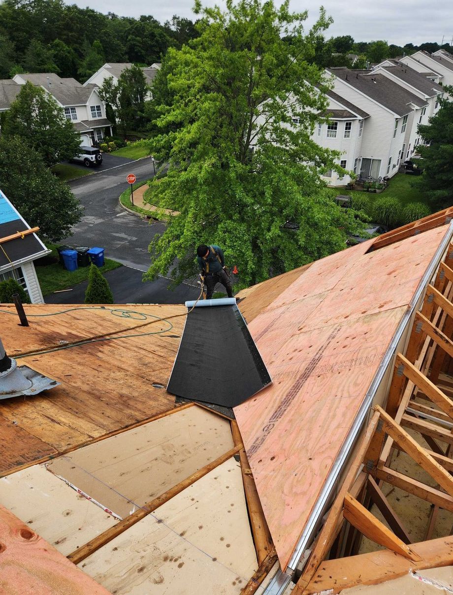 A worker installs plywood sheeting on a residential roof frame near a suburban neighborhood on an overcast day.