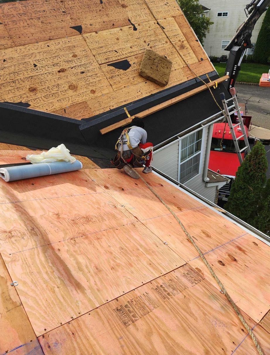 A worker in a safety harness kneels on a partially re-sheeted plywood roof while installing new roofing materials.