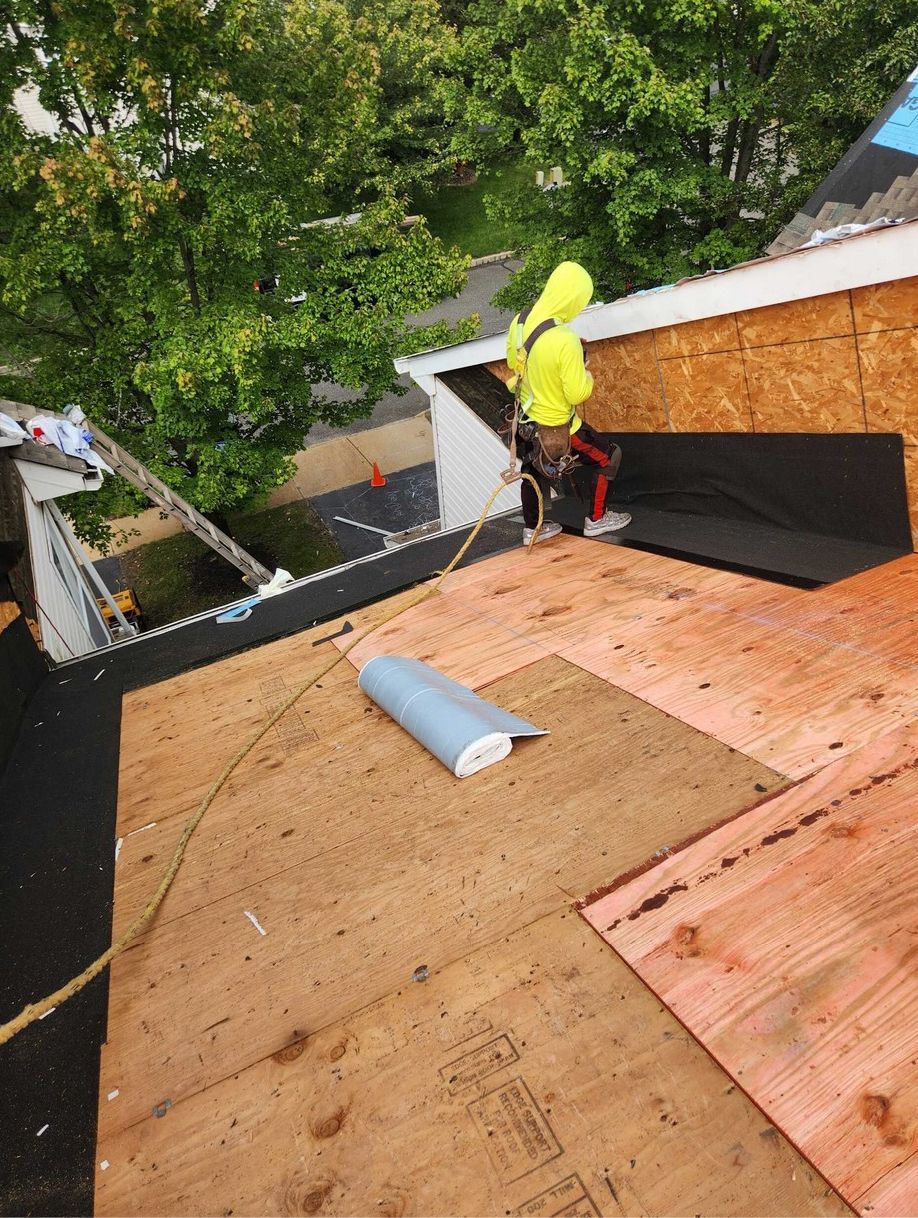 A worker in a bright yellow hoodie and safety harness installs roofing materials on a wooden residential roof.
