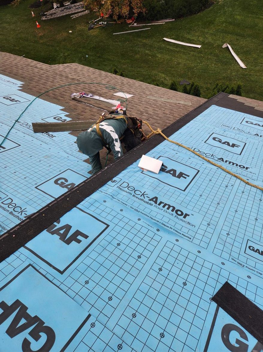 A roofer in a safety harness works on a residential roof covered in blue GAF Deck-Armor underlayment.