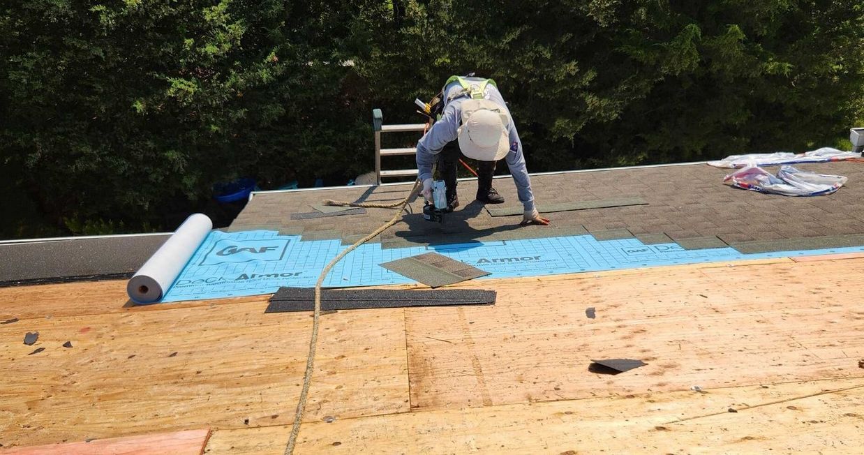 A person wearing a safety harness installs blue roofing underlayment onto a wooden roof deck next to a rolled material.
