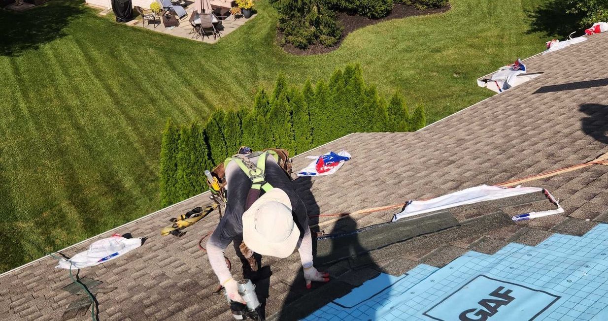A worker wearing a safety harness and hat installs shingles on a sloped roof with blue underlayment, viewed from above.