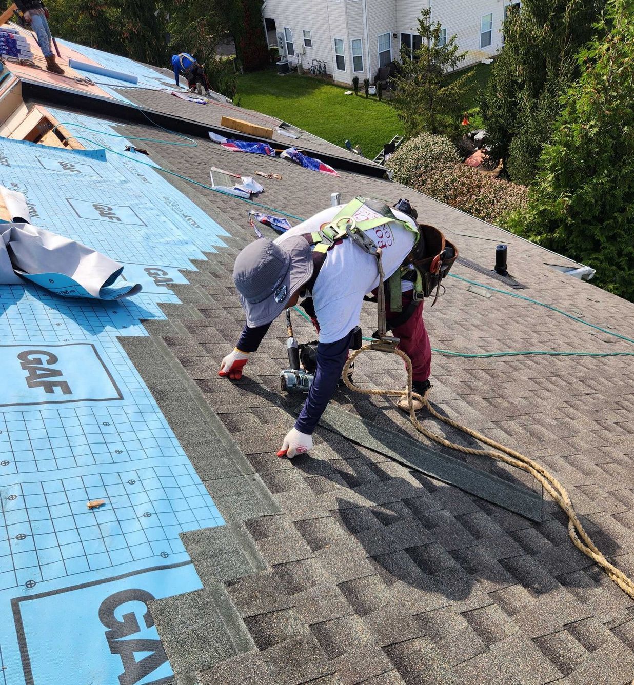 A roofer wearing a harness and sun hat works on a residential roof, installing shingles over blue underlayment.
