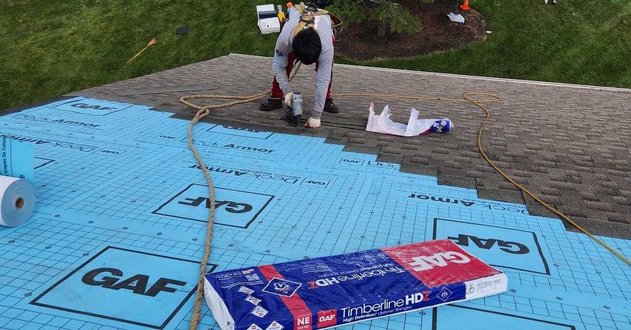 A worker installs shingles on a residential roof covered in blue GAF underlayment.