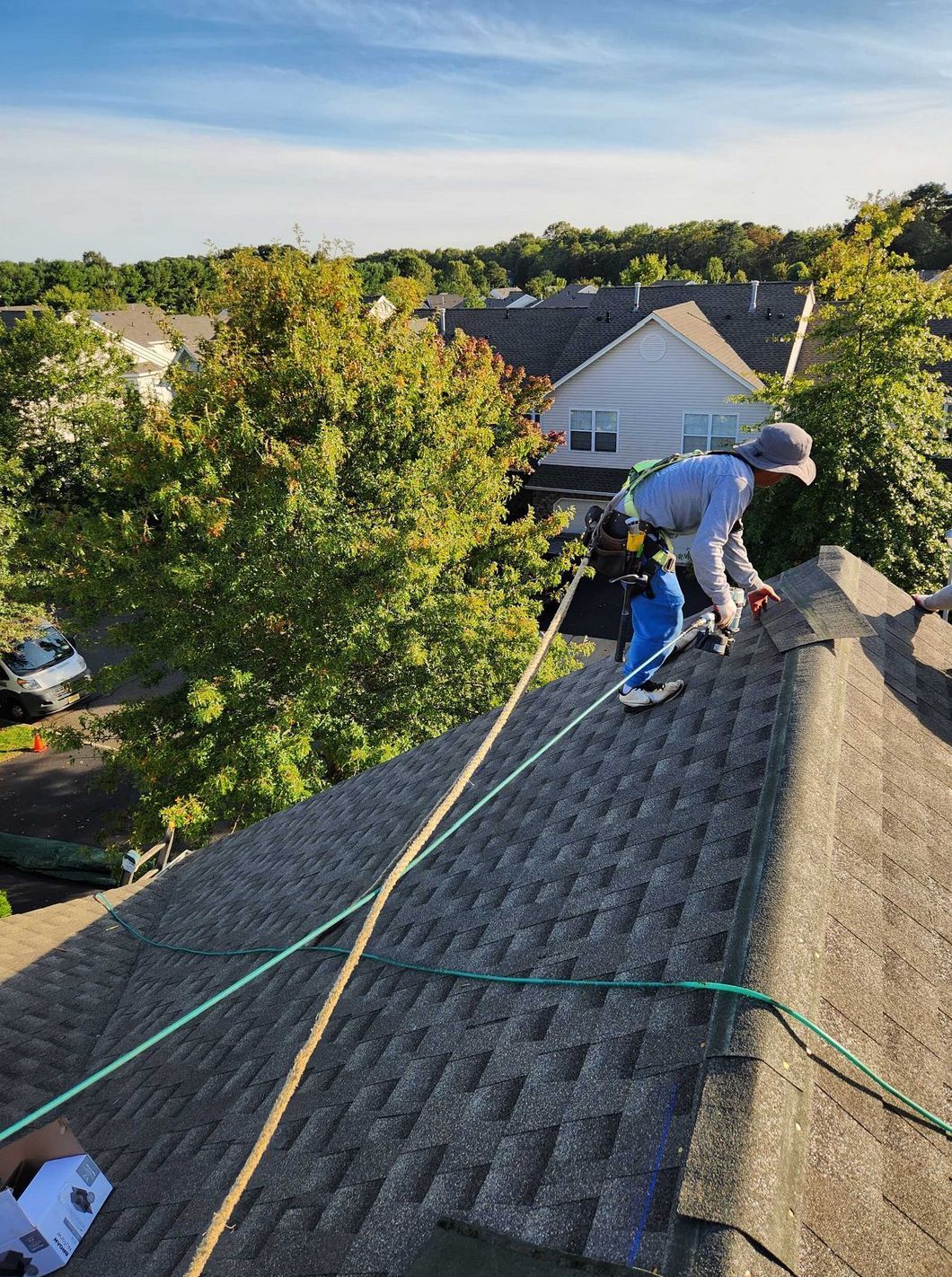A worker in safety gear, tethered by a rope, inspects a residential shingled roof under a clear blue sky.