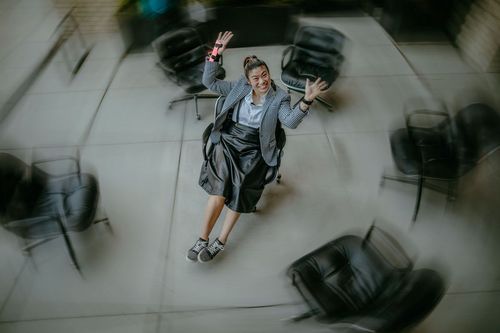 Woman in business attire playfully spins on a rolling chair in an office; blurry motion effect.