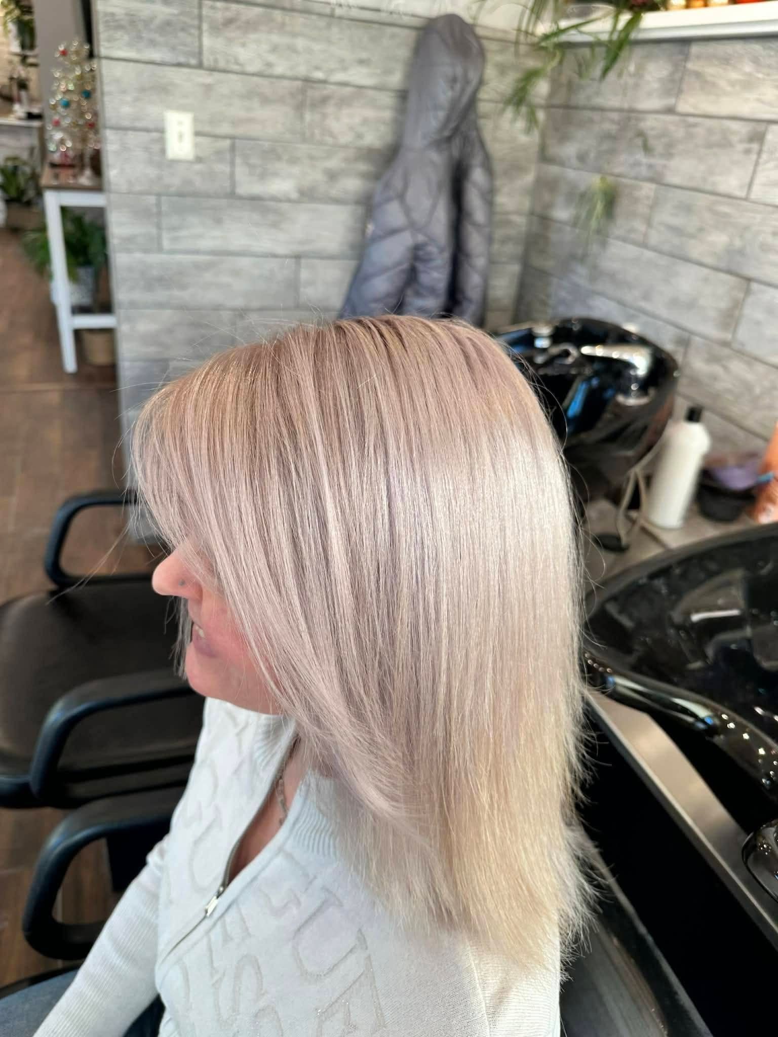 Woman with light pink hair in a salon chair, facing left. White sweater, brick-patterned wall, sink visible.