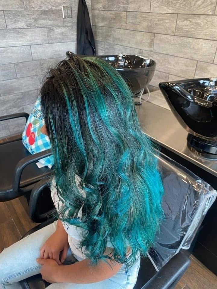 Woman with long, wavy, teal-colored hair in a salon chair. Black roots blend into the bright teal.