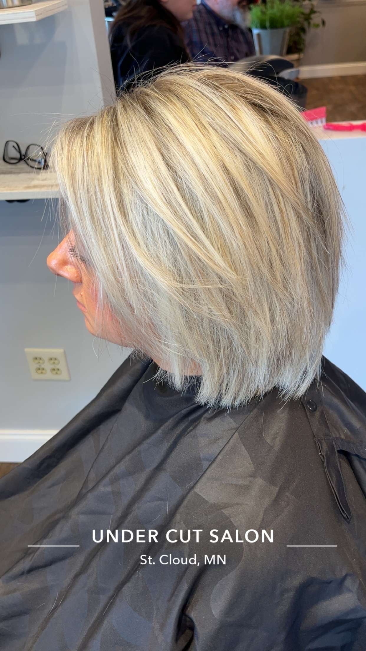 Woman with short, layered, blonde hair at a salon. She wears a black salon cape, profile view.