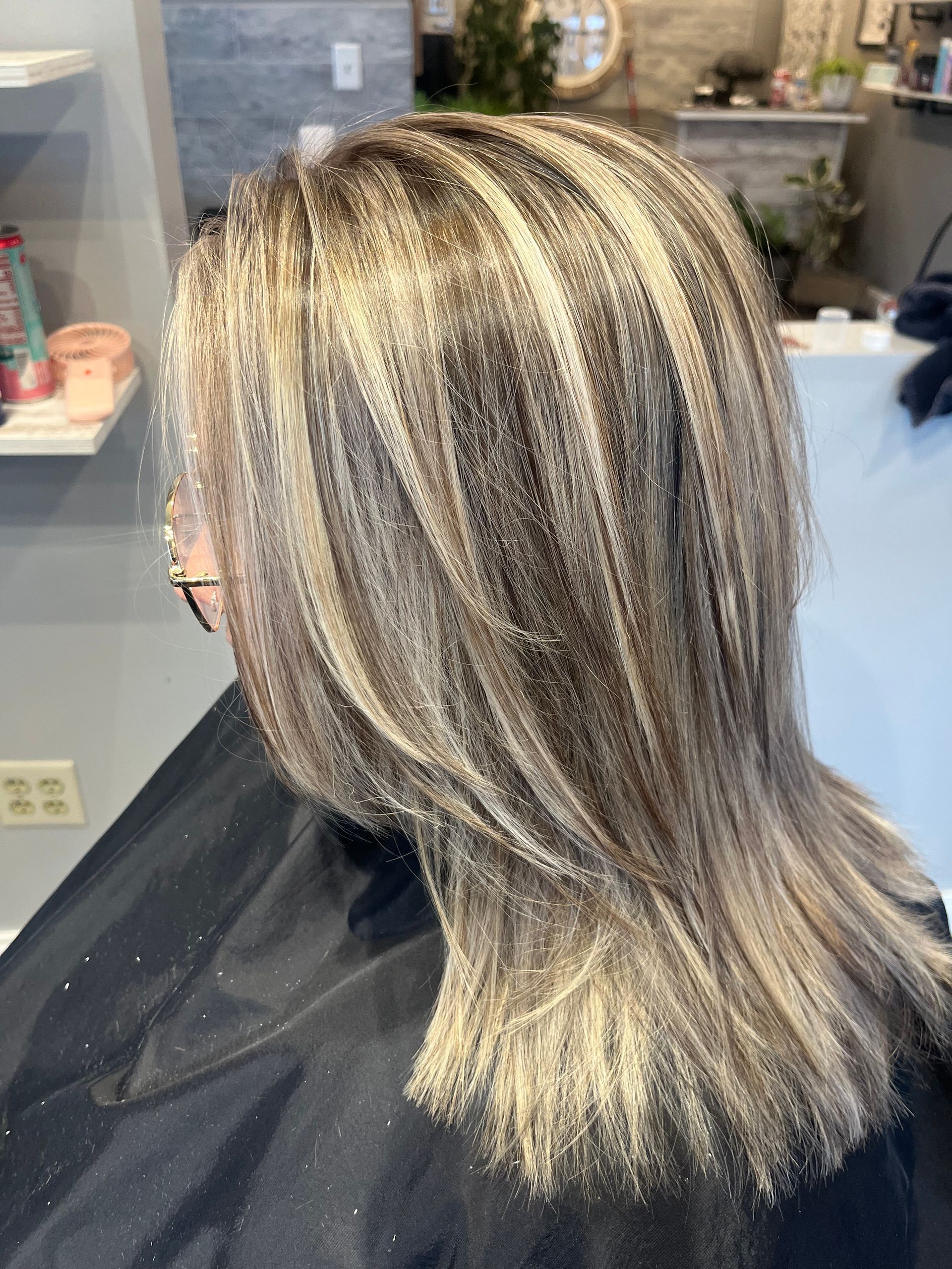 Woman with layered, blonde highlighted hair at a salon, wearing a black cape. 