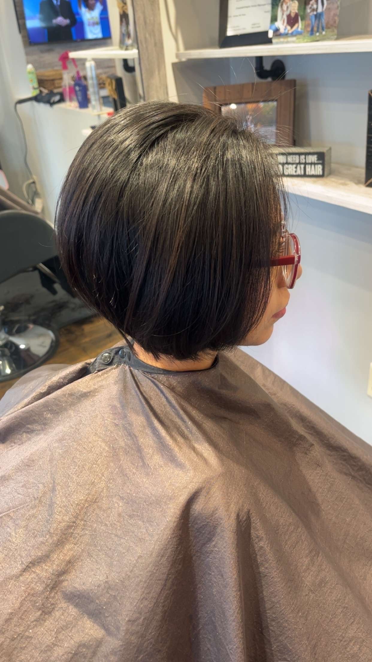 Person with short, dark hair in a salon chair, wearing a protective cape. Hair is styled in a rounded bob. 