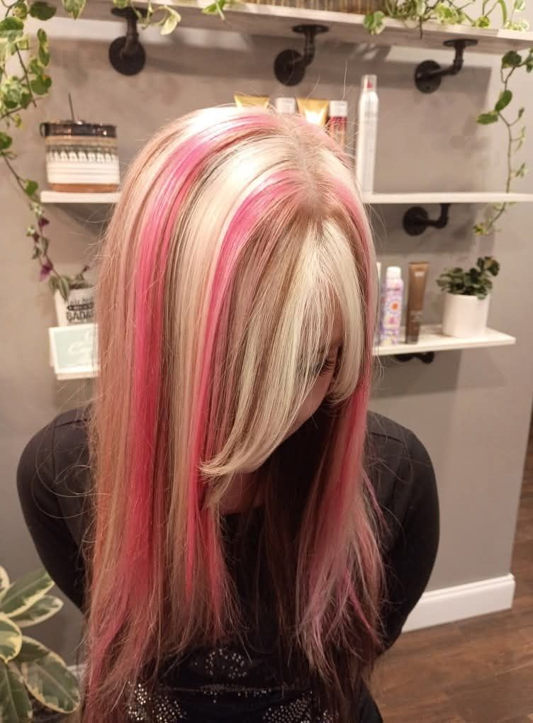 Woman with long blonde hair, streaks of pink. In a salon setting, shelving in the background. 