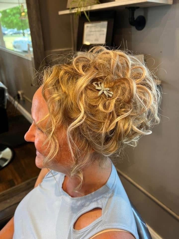 Woman with curly blonde hair styled in an updo, wearing a light blue shirt, in a salon.