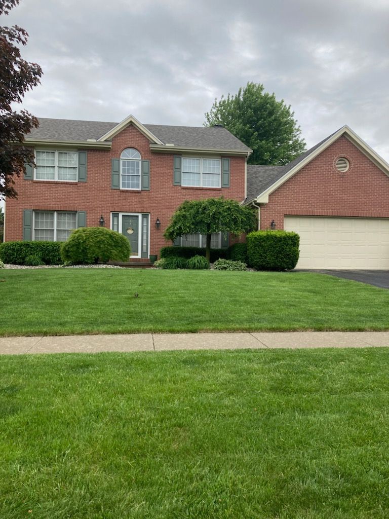 A two-story red brick house with a white garage, green shutters, and well-manicured bushes on a grassy lawn.