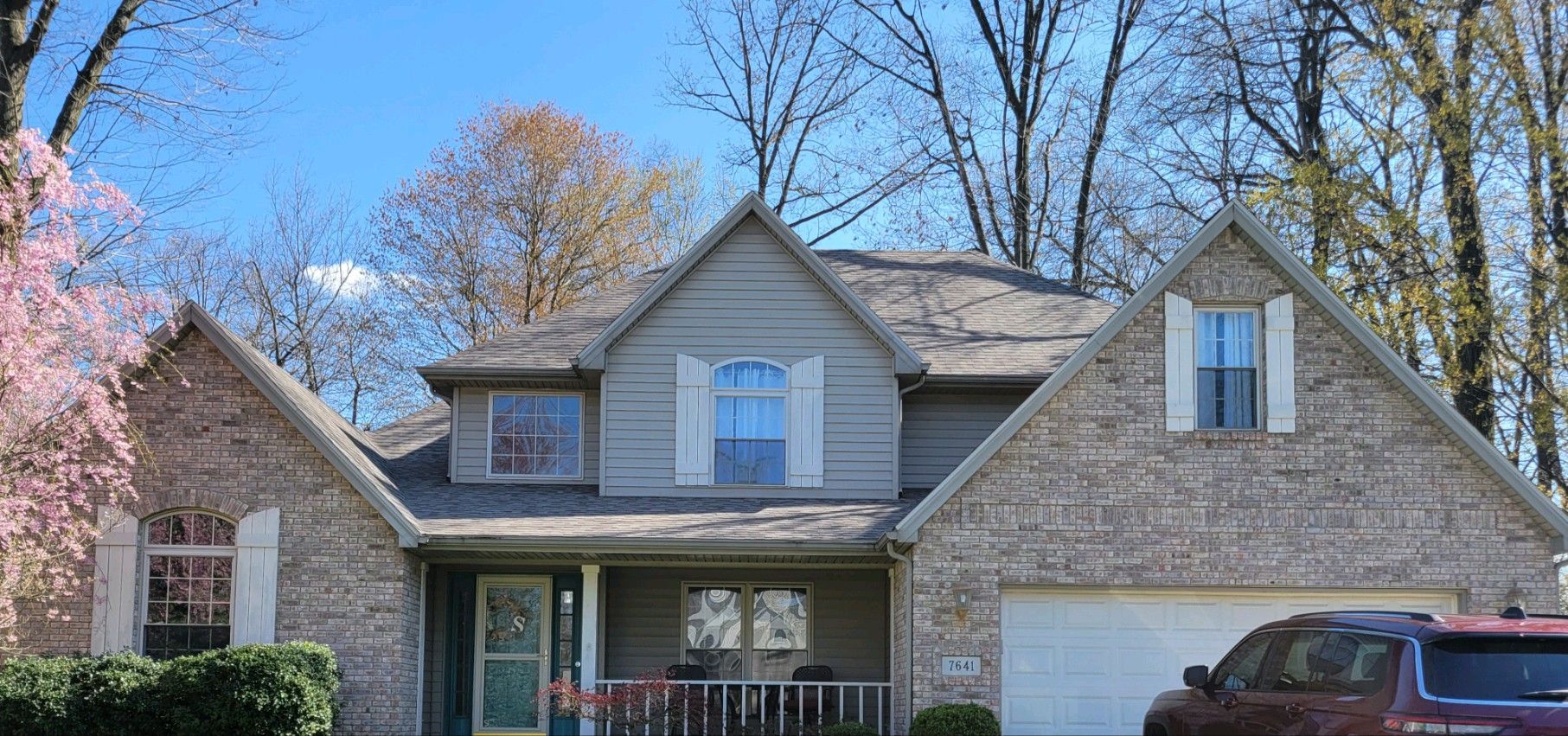 A two-story tan brick house with a front porch, gabled roof, and white-trimmed windows under a clear blue sky.