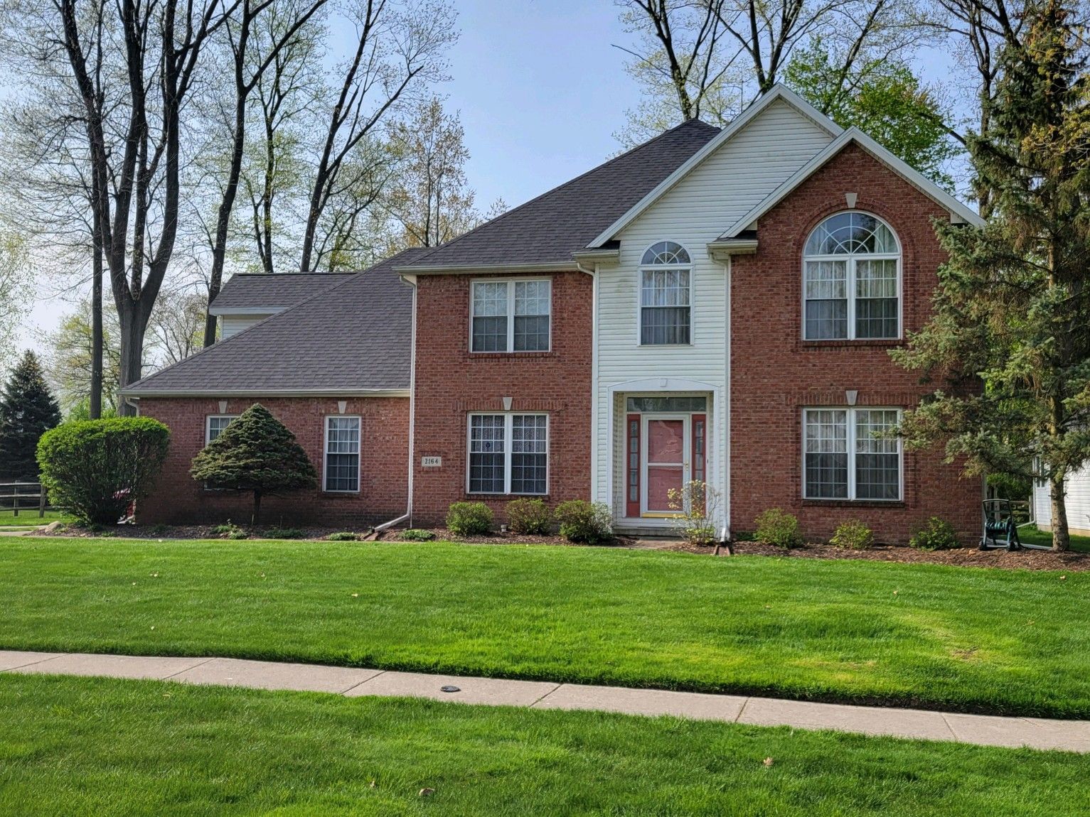 A two-story, red brick house with a white front entryway and gabled roof, set behind a large green lawn on a sunny day.