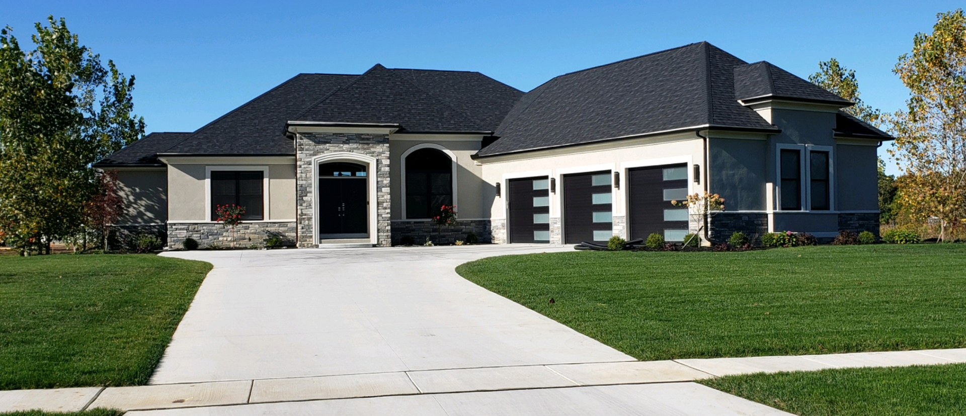 A tan, single-story house with a black roof, stone accents, and a three-car garage, set against a green lawn and blue sky.