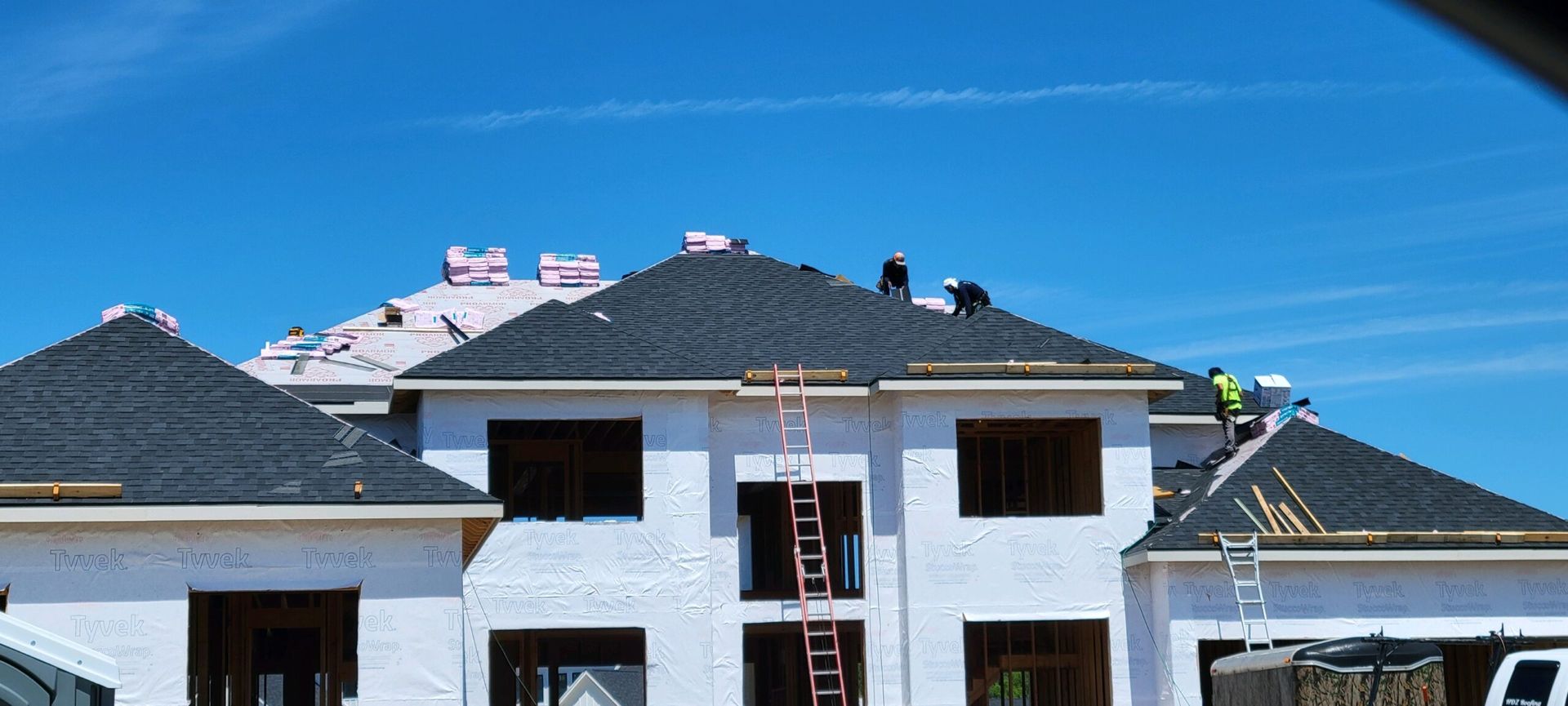 Construction workers install black shingles on the roof of a large, unfinished white house under a clear blue sky.