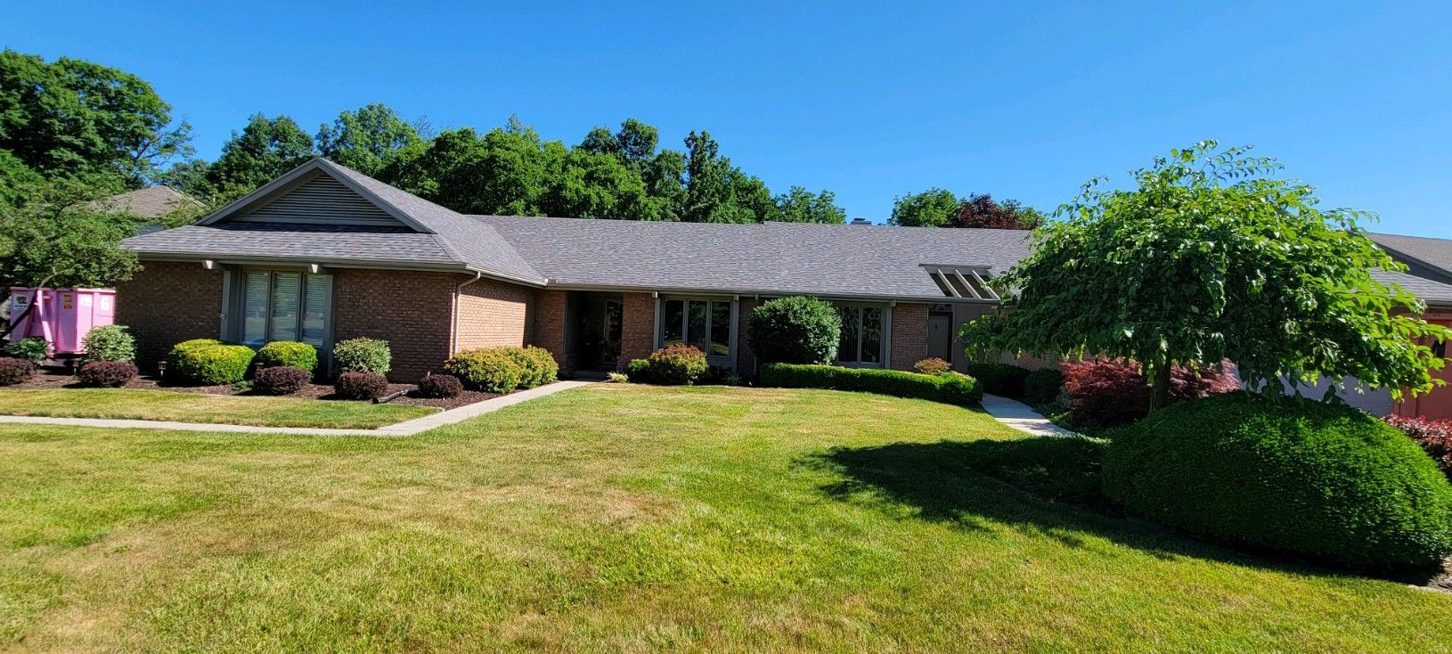A single-story brick house with a gray shingled roof, surrounded by a green lawn, shrubs, and trees under a clear blue sky.