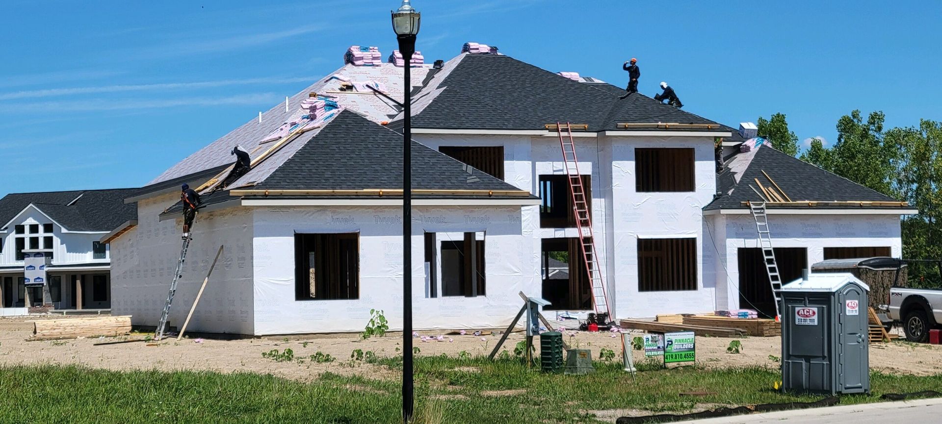 Workers install dark shingles on the roof of a large, white, under-construction house on a sunny day.