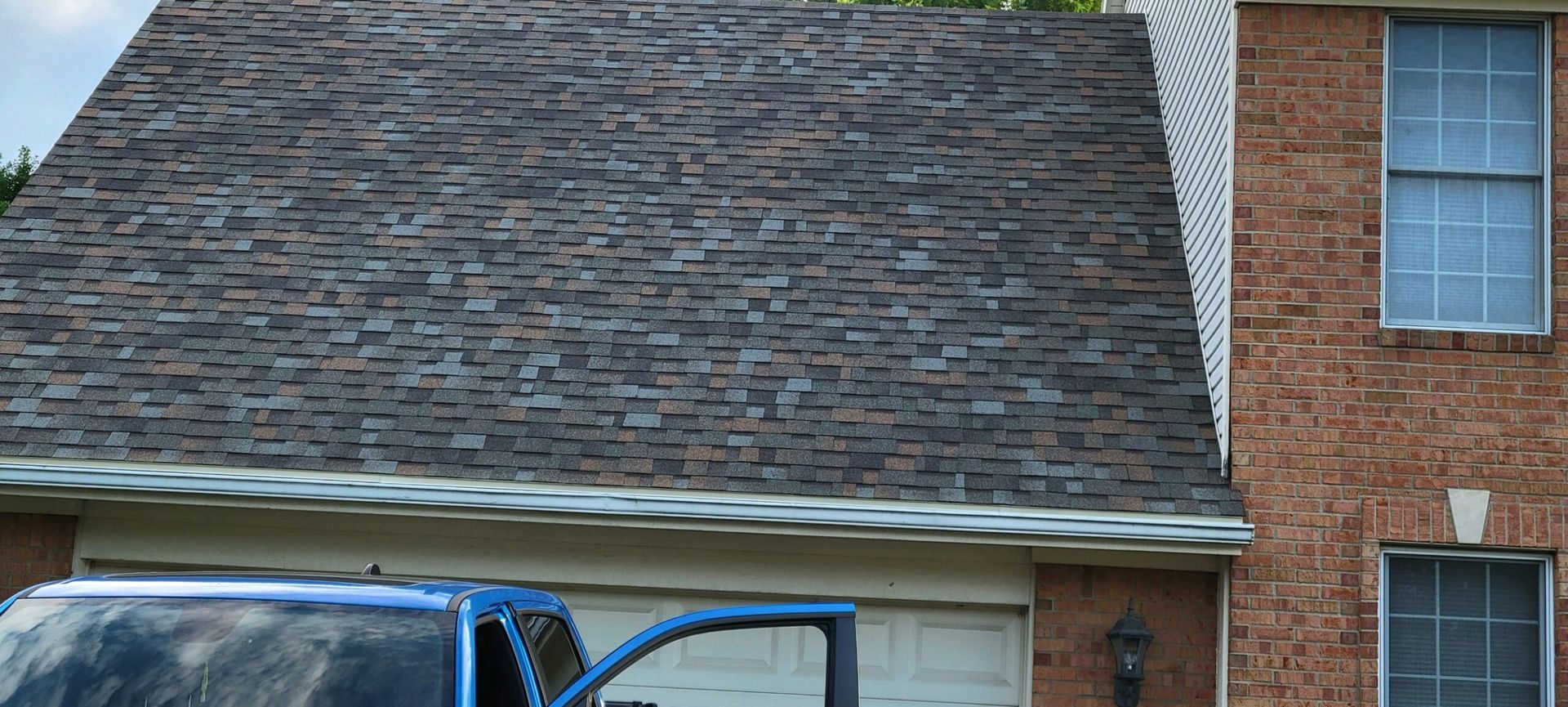 A blue pickup truck parked in front of a brick house with a dark shingled roof and white siding.