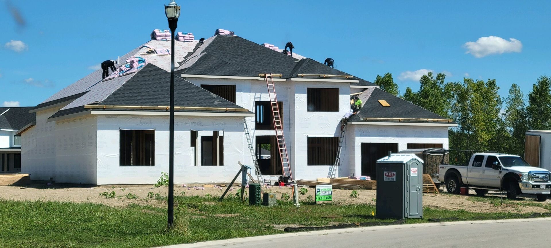 A multi-story house under construction with a dark shingled roof, white exterior walls, a porta-potty, and a pickup truck.