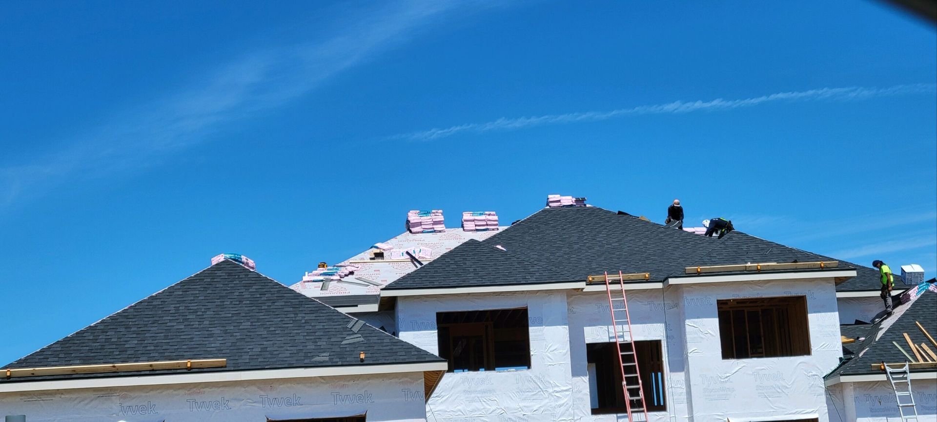 Construction workers install shingles on the roof of a house under a clear blue sky.
