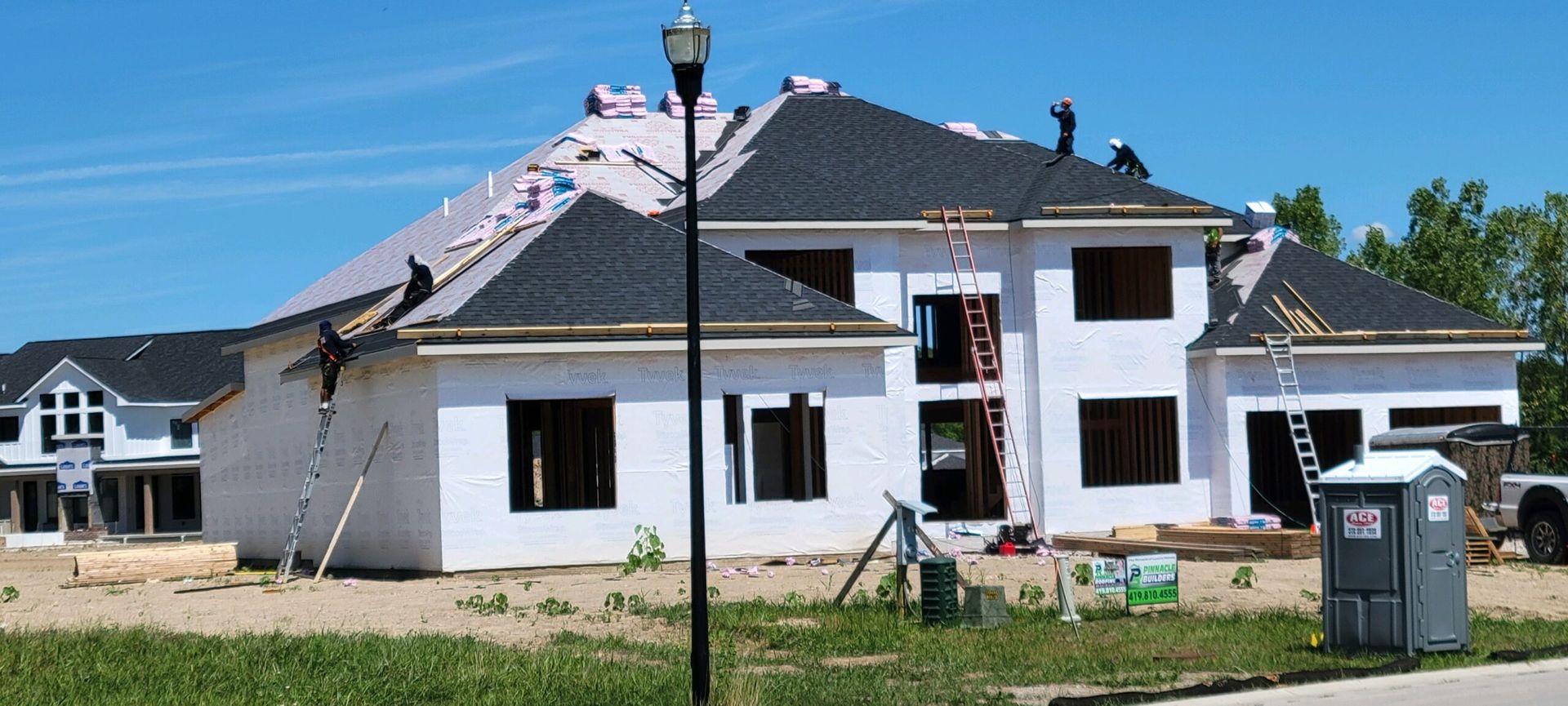 Workers install a dark shingled roof on a white, two-story house under construction on a sunny day.