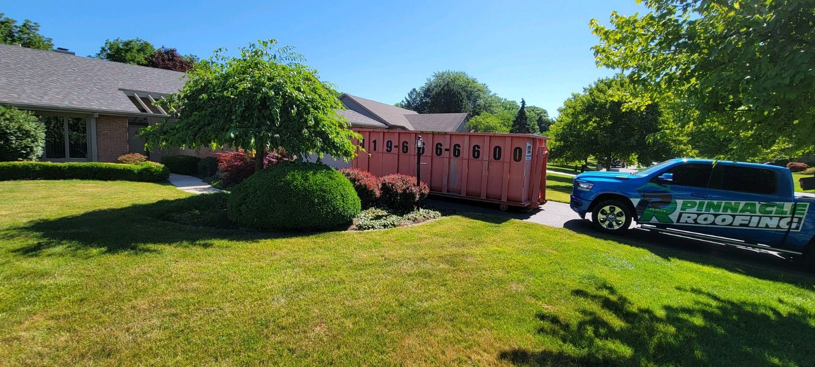 A blue service truck parked on a sunny lawn next to a large, empty red dumpster in front of a residential house.