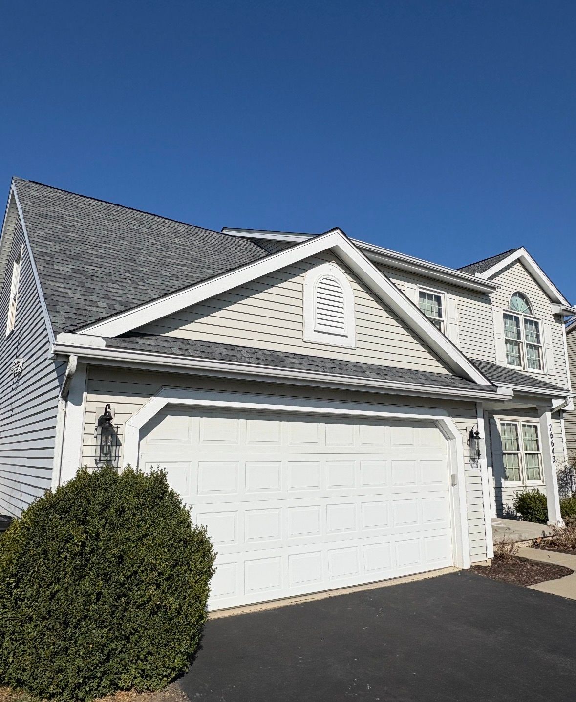 Suburban house exterior with white garage doors, gray siding, and a dark driveway under a clear blue sky