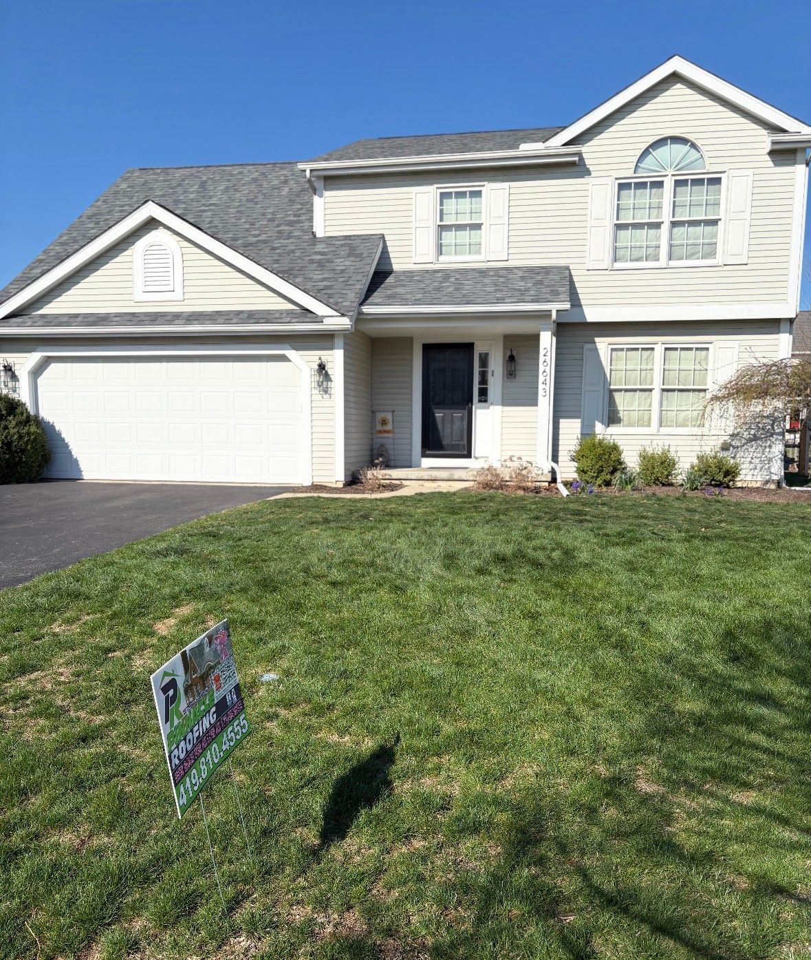 Suburban two-story white house with garage and front lawn under bright sunlight, real estate sign in yard