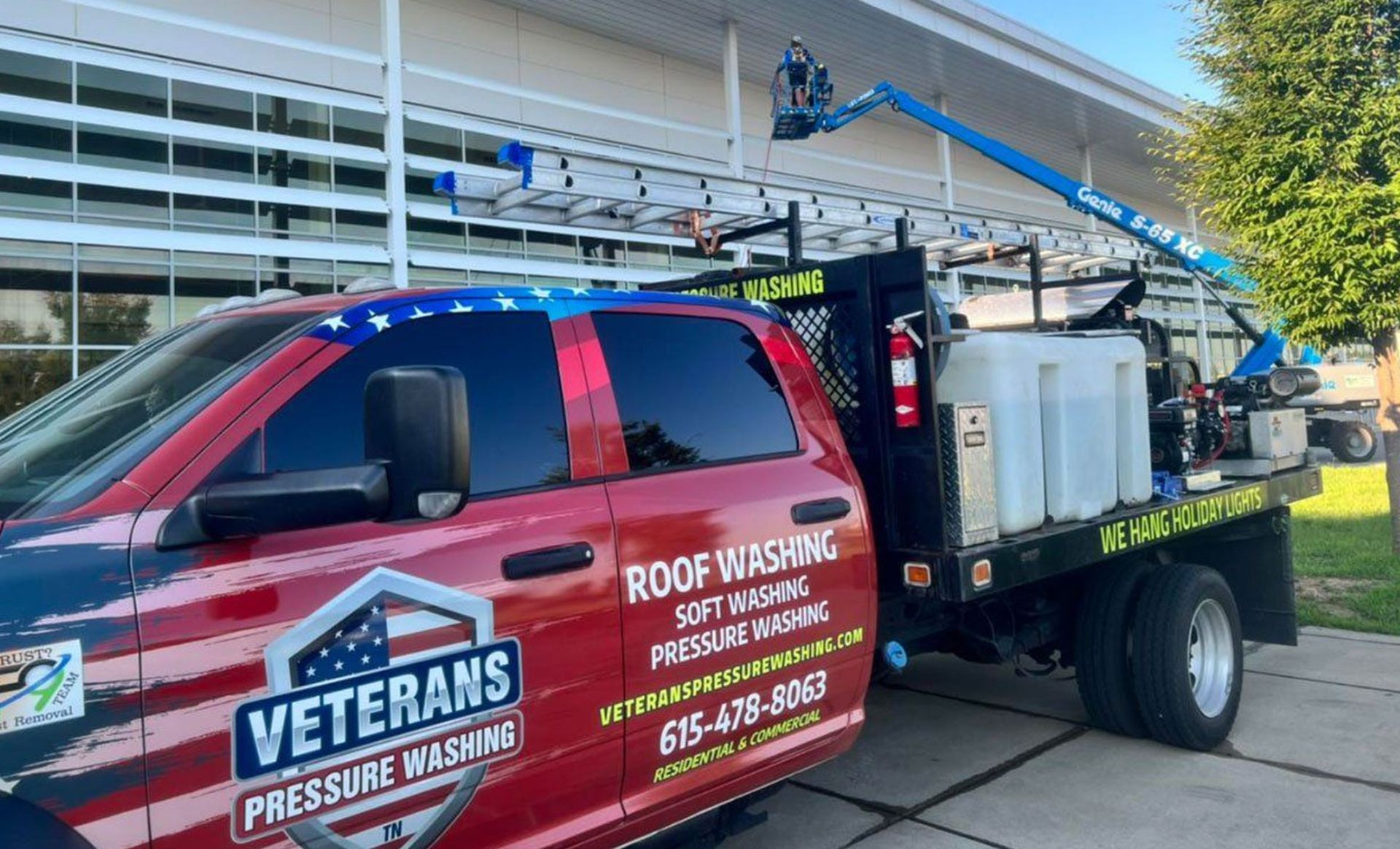 Truck with boom lift, cleaning building exterior. Veteran-owned pressure washing service. Red, white, blue color scheme.